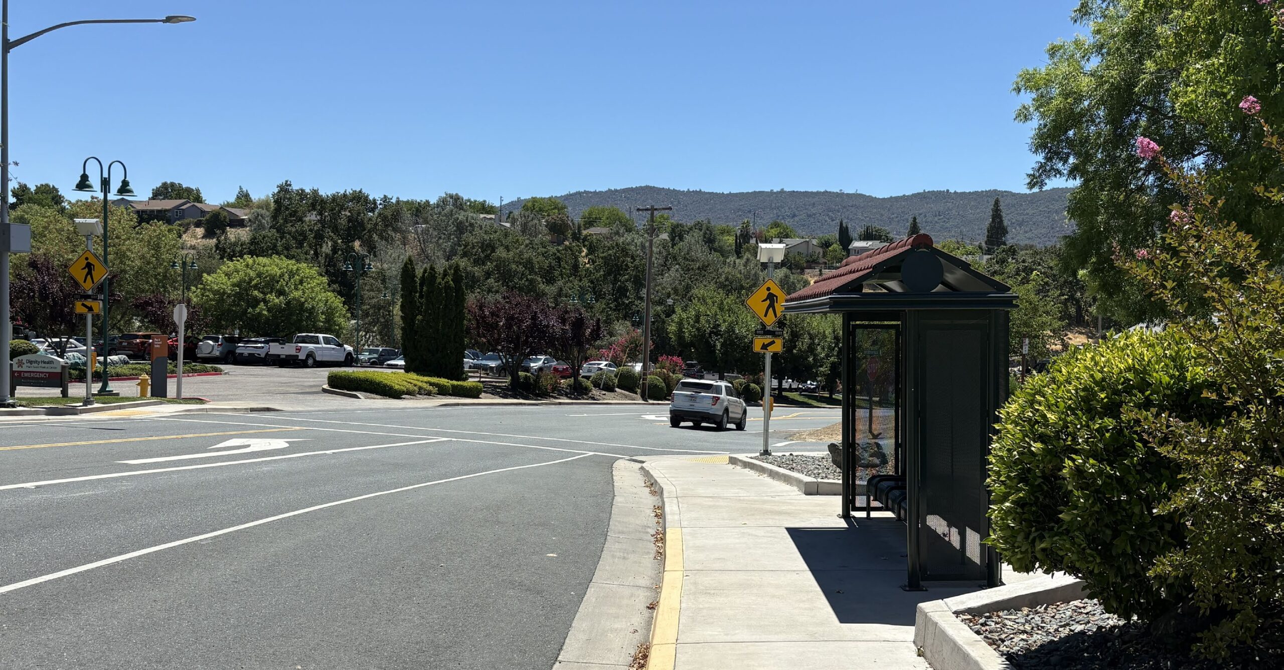 A quiet street with a bus stop shelter on the right, a car making a turn, trees, parked cars, and hills in the background under a clear blue sky.