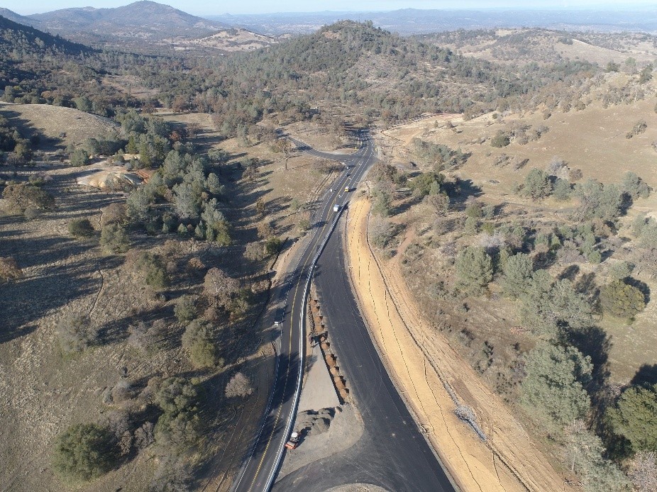Aerial view of a partially constructed road winding through a hilly, rural landscape with sparse trees and mountains in the background.