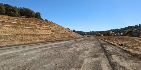 A wide, newly graded dirt road curves alongside a terraced, dry hillside under a clear blue sky.