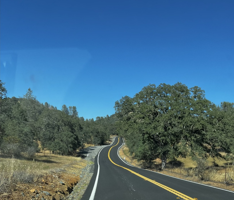 A two-lane road with yellow dividing lines curves through a dry, wooded landscape under a clear blue sky.