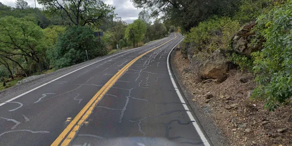 A two-lane road with yellow centerlines winds uphill through a green, wooded area under a cloudy sky. Cracks in the asphalt have been filled with sealant.