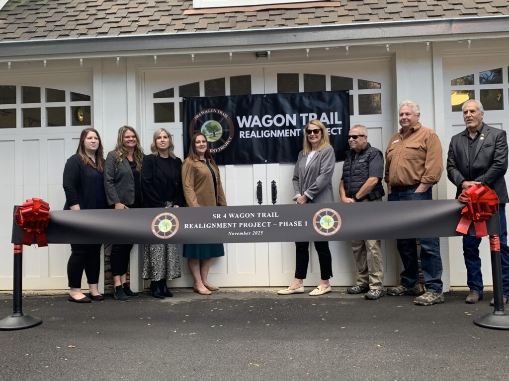 Eight people stand in front of a building and banner for the Wagon Trail Realignment Project behind a ribbon labeled “SR 4 Wagon Trail Realignment Project – Phase 1, November 2023.”.
