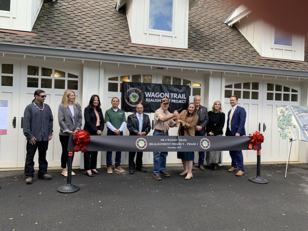 A group of people stand in front of a building, cutting a ribbon labeled “Wagon Trail Realignment Project – Phase II” for a ceremonial event.
